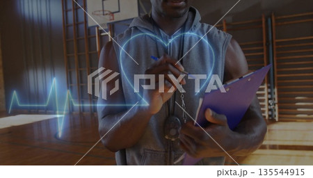 Observing fitness coach in hoodie monitoring heartbeat on gym court with whistle and clipboard Observing fitness coach in hoodie monitoring heartbeat on gym court with whistle and clipboard 135544915