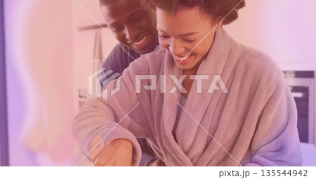 Bathrobe wearing woman cooking with smiling partner in kitchen, with wooden spatula and mixing bowl Bathrobe wearing woman cooking with smiling partner in kitchen, with wooden spatula and mixing bowl 135544942