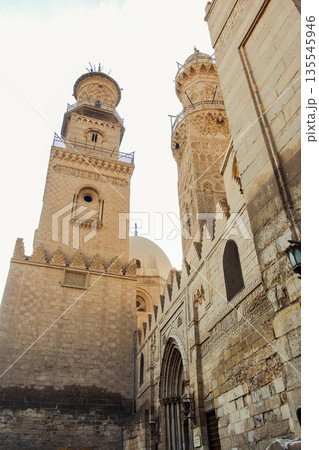 Historic Qalawun Complex in Cairo, featuring intricate architecture, tall minarets, and domes under a blue sky with clouds. A significant cultural landmark on Al-Muizz Street. 135545946