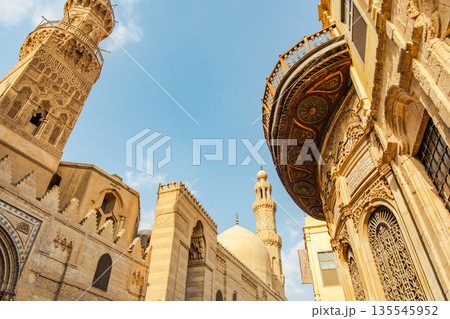 Historic Qalawun Complex in Cairo, featuring intricate architecture, tall minarets, and domes under a blue sky with clouds. A significant cultural landmark on Al-Muizz Street. 135545952