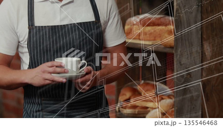 Barista wearing striped apron holding white ceramic cup and saucer in bakery, with bread loaves 135546468