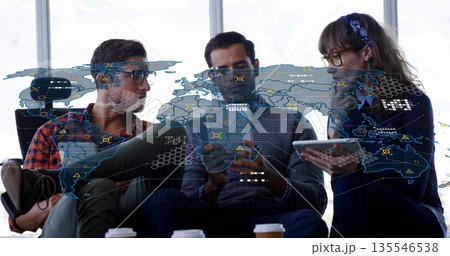 Three professionals discussing data sitting in office lounge, with devices, coffee and map overlay 135546538