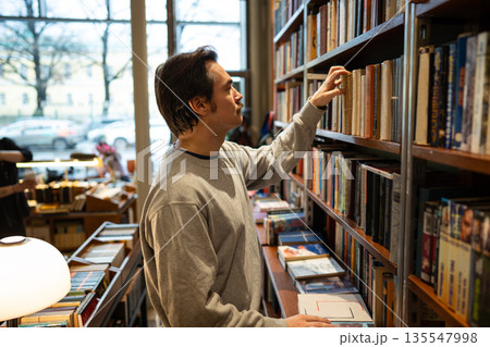 Curious focused man booklover picking hardcover book from bookshelf in modern bookstore library. Curious focused man booklover picking hardcover book from bookshelf in modern bookstore library. 135547998