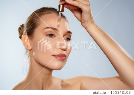 Woman applying serum to her face with a dropper in a well-lit setting Woman applying serum to her face with a dropper in a well-lit setting 135548108