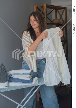 Morning home ritual with brunette carefully preparing clothes for work day holding an iron in hand 135548472