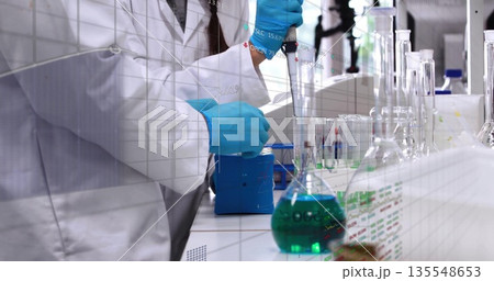 Using micropipette scientist wearing lab coat transferring green solution into flask at lab bench Using micropipette scientist wearing lab coat transferring green solution into flask at lab bench 135548653