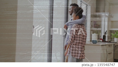 Embracing couple gazing through sliding glass door in kitchen with mug plant utensils, copy space Embracing couple gazing through sliding glass door in kitchen with mug plant utensils, copy space 135548747