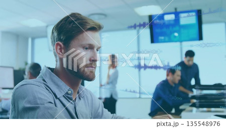 Focusing bearded man in button-down shirt typing on computer in open-plan office with data display 135548976