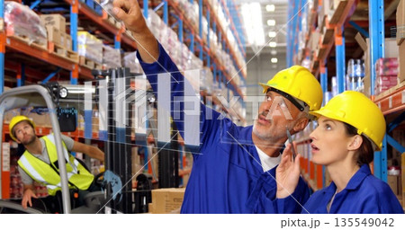 Pointing staff in blue coveralls and hard hats standing in warehouse, with forklift, chart overlay 135549042