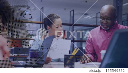 Man in pink shirt pointing with woman in blue top listening at office desk, digital overlay 135549832