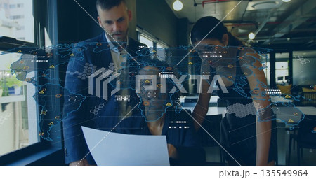 Colleagues reviewing printed report wearing business attire in open office, with world map overlay 135549964