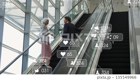 Talking business colleagues climbing escalator holding rail in glass atrium, with social icons 135549966