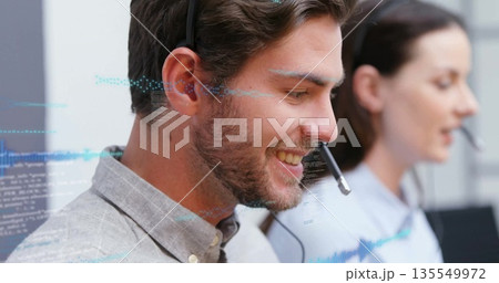 Smiling male call center agent speaking into headset at office desk, with holographic code overlays 135549972