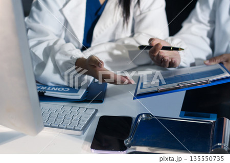 Medical team having a meeting with doctors in white lab coats and surgical scrubs seated at a table discussing a patients working online using computers in the medical industry. Medical team having a meeting with doctors in white lab coats and surgical scrubs seated at a table discussing a patients working online using computers in the medical industry. 135550735