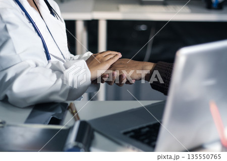 Senior patient lying in hospital bed receiving comforting touch from young female nurse, hands gently clasped together, medical care setting visible 135550765