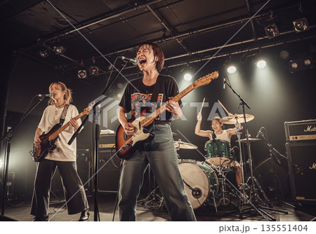 ライブハウスで熱唱する女性ボーカルギターとバンドメンバーの激しい演奏 ライブハウスで熱唱する女性ボーカルギターとバンドメンバーの激しい演奏 135551404