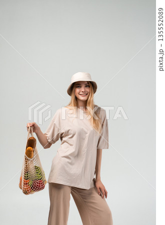 Smiling young woman carrying reusable grocery bag with fresh produce, minimal eco friendly shopping lifestyle on light background Smiling young woman carrying reusable grocery bag with fresh produce, minimal eco friendly shopping lifestyle on light background 135552089