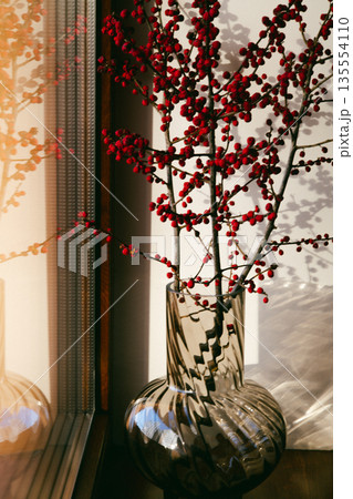 Minimalist interior still life featuring a transparent glass vase with red berry branches placed on a windowsill. A play of shadows and light on wall. 135554110