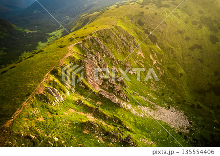 Breathtaking aerial view of green, rugged mountain peaks under dramatic sky. Scene captures contrast between vibrant landscape and moody clouds, with sunlight breaking through. Carpathians, Chornogora Breathtaking aerial view of green, rugged mountain peaks under dramatic sky. Scene captures contrast between vibrant landscape and moody clouds, with sunlight breaking through. Carpathians, Chornogora 135554406