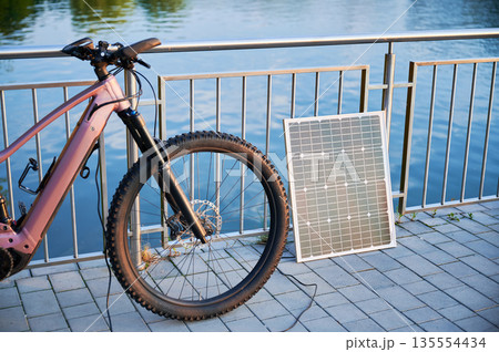 Pink electric mountain bike parked beside lakeside railing, connected to solar panel for charging. Bicycle's sleek design and rugged tires highlighted, with tranquil lake and golden light. 135554434