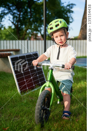 Young boy sits on green balance bike next to solar panel on grass. Kid wearing green helmet, looks at camera. Background features fence, trees, and bench. Concept of green energy generation. 135554438