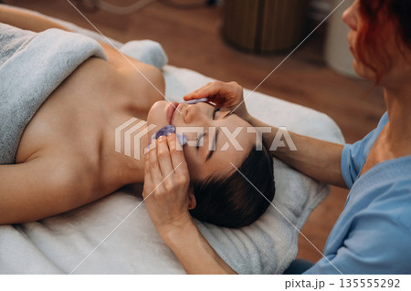 Cosmetology, face skin care. Relaxed young woman getting massage by therapist in spa salon Cosmetology, face skin care. Relaxed young woman getting massage by therapist in spa salon 135555292