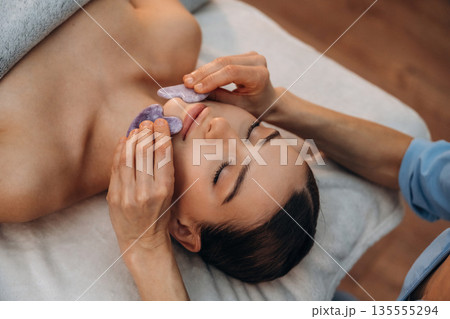 Cosmetology, face skin care. Relaxed young woman getting massage by therapist in spa salon 135555294