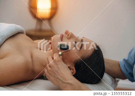 Black roller massager. Relaxed young woman getting massage by therapist in spa salon 135555312