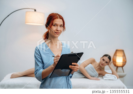 Holding notepad. Female masseur near woman lying down in spa salon Holding notepad. Female masseur near woman lying down in spa salon 135555531
