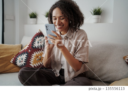 Woman With Bright Smile Reviews Playful Message On Her Smartphone Surrounded By Cozy Decor And Cushions 135556113