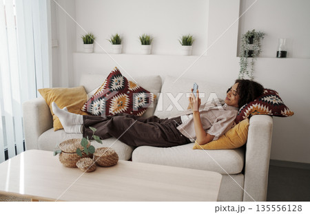 Woman Lying Comfortably On Sofa, Serene Lady Reclined On Couch While Scrolling Through Her Device Woman Lying Comfortably On Sofa, Serene Lady Reclined On Couch While Scrolling Through Her Device 135556128