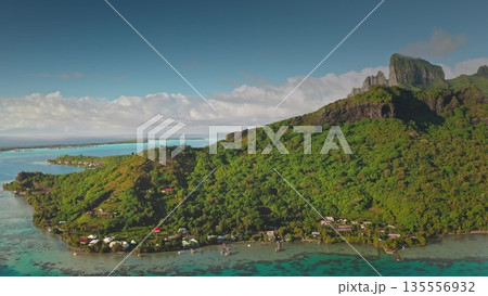 Aerial panoramic view revealing pristine Bora Bora coastline with verdant tropical vegetation. Crystal-clear turquoise lagoon. Traditional houses. Wooden piers nestled in French Polynesian landscape 135556932