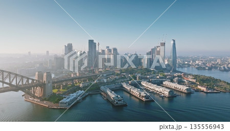 Australia, Sydney: Aerial view of Sydney Harbour Bridge and city skylines skyscrapers reflecting in water at misty sunrise, Barangaroo Reserve and wharves in foreground. Drone flight panorama 135556943