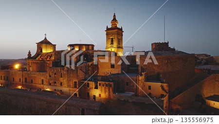 Malta, Comino island: Warm night lights illuminating the Cittadella in Gozo, Malta, highlight the medieval architecture of the fortified city at dusk. Aerial drone flight panorama Malta, Comino island: Warm night lights illuminating the Cittadella in Gozo, Malta, highlight the medieval architecture of the fortified city at dusk. Aerial drone flight panorama 135556975