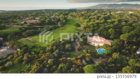 Portugal, Lisbon: Aerial view golf course in Cascais with lush green meadow ad trees landscape, luxury villas houses, mountain and ocean in background. Beautiful sunset scene. Drone flight Portugal, Lisbon: Aerial view golf course in Cascais with lush green meadow ad trees landscape, luxury villas houses, mountain and ocean in background. Beautiful sunset scene. Drone flight 135556979