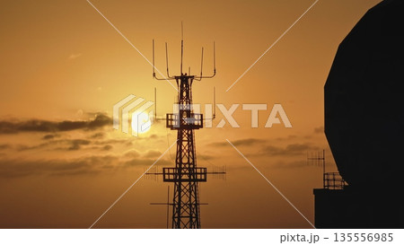 Telecommunication tower and parabolic antenna silhouette against vibrant orange sunset sky, essence of communication and technology in a breathtaking evening landscape. Aerial drone flight panorama Telecommunication tower and parabolic antenna silhouette against vibrant orange sunset sky, essence of communication and technology in a breathtaking evening landscape. Aerial drone flight panorama 135556985