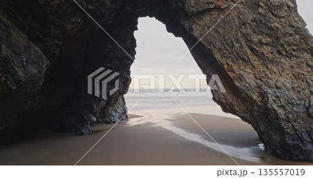 Portugal, Lisbon: rock arch at Praia da Adraga beach framing the Atlantic ocean, waves breaking on wet sand shore under cloudy sky. Low angle drone flight in mountain cliff hole. Nature background Portugal, Lisbon: rock arch at Praia da Adraga beach framing the Atlantic ocean, waves breaking on wet sand shore under cloudy sky. Low angle drone flight in mountain cliff hole. Nature background 135557019
