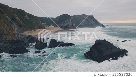 Portugal, Lisbon: Praia da Adraga rugged Atlantic coast, powerful waves crashing on cliffs rock, sea under an overcast sky. Tourist people walk sandy beach, enjoy dramatic scene. Aerial drone flight Portugal, Lisbon: Praia da Adraga rugged Atlantic coast, powerful waves crashing on cliffs rock, sea under an overcast sky. Tourist people walk sandy beach, enjoy dramatic scene. Aerial drone flight 135557026