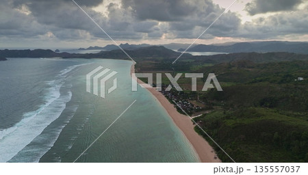 Aerial view capturing the expansive Pantai Tomang Omang beach coastline, with gentle ocean waves breaking on its sandy shore under a cloudy sky, showing a small village nestled by the green hills Aerial view capturing the expansive Pantai Tomang Omang beach coastline, with gentle ocean waves breaking on its sandy shore under a cloudy sky, showing a small village nestled by the green hills 135557037