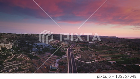 Malta: highway road with driving cars crossing the evening desert island at sunset, with a colorful pink cloudy sky and cultivated fields. Dramatic nature landscape. Aerial view drone flight 135557049
