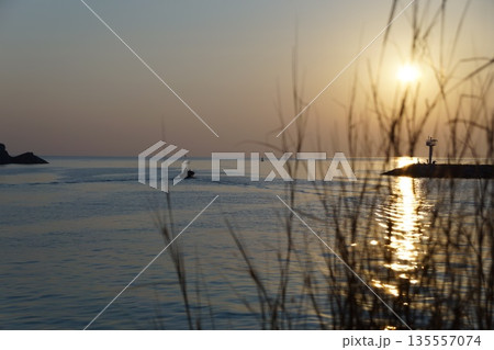 Dawn of the golden sun.The golden sun rises from behind the blue sky on a stone pier and lighthouse, against a backdrop of trees and grass. Dawn of the golden sun.The golden sun rises from behind the blue sky on a stone pier and lighthouse, against a backdrop of trees and grass. 135557074