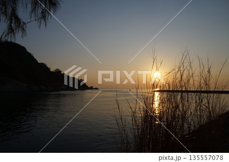 The golden sun rises from behind the blue sky on a stone pier and lighthouse, against a backdrop of trees and grass. 135557078