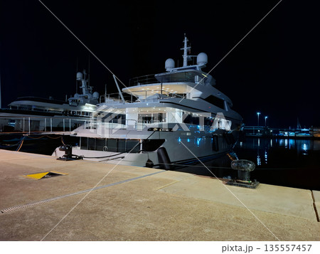 A large yacht is docked at a marina in Spain as night falls. The yacht stands out with lights shining on its structure while calm waters reflect the scene. 135557457