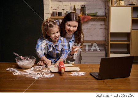 Mother and daughter smiling while preparing dough at home table. Mother and daughter smiling while preparing dough at home table. 135558250