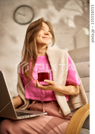 Smiling woman relaxing with red juice while using laptop at cozy home workspace. 135558555