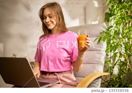 Young woman working on laptop while holding glass of fresh orange juice at home. 135558556