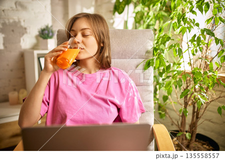 Smiling woman drinking fresh orange juice while working from home. Smiling woman drinking fresh orange juice while working from home. 135558557