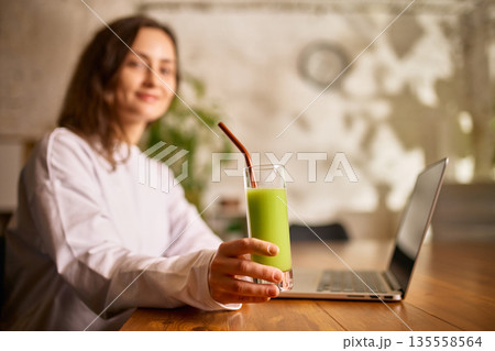 Woman holding green smoothie glass while working on laptop at home. 135558564