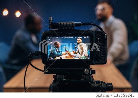 Close up view of camera that is filming. Two radio presenters recording an audio broadcast in a studio 135559243