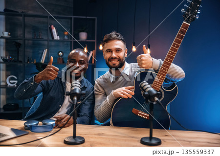 Posing, holding acoustic guitar. Two radio presenters recording an audio broadcast in a studio Posing, holding acoustic guitar. Two radio presenters recording an audio broadcast in a studio 135559337